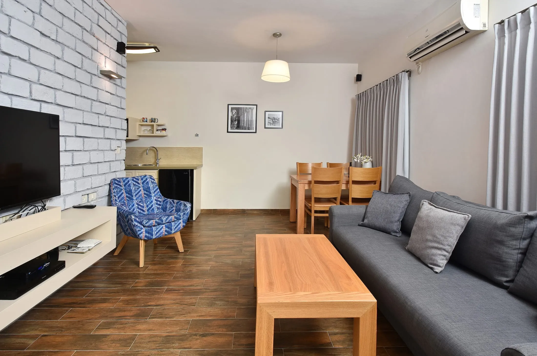 Suite living room with grey sofa, brick accent wall, dining table and kitchenette