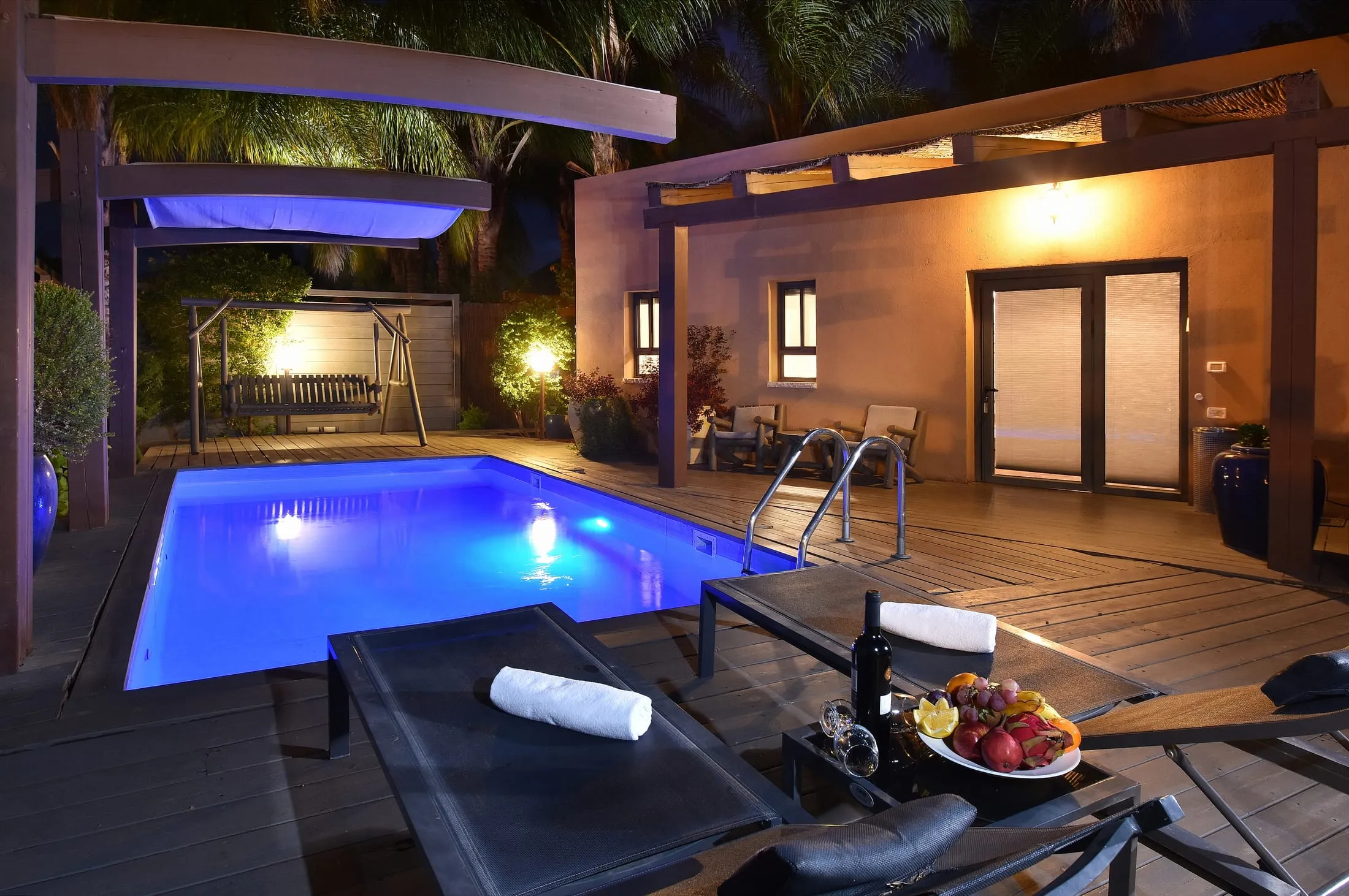 Private pool of a luxury suite lit blue at night, with loungers, wine and fruit platter on the deck